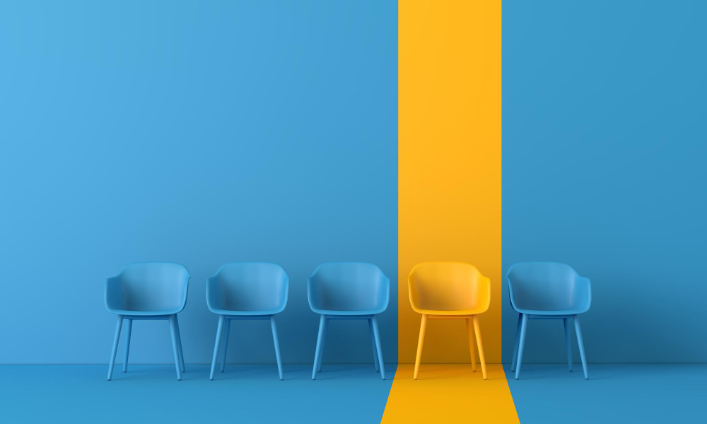 Cinco cadeiras em um fundo azul, quatro delas são azuis da cor do fundo, e uma delas é amarela, se destacando.